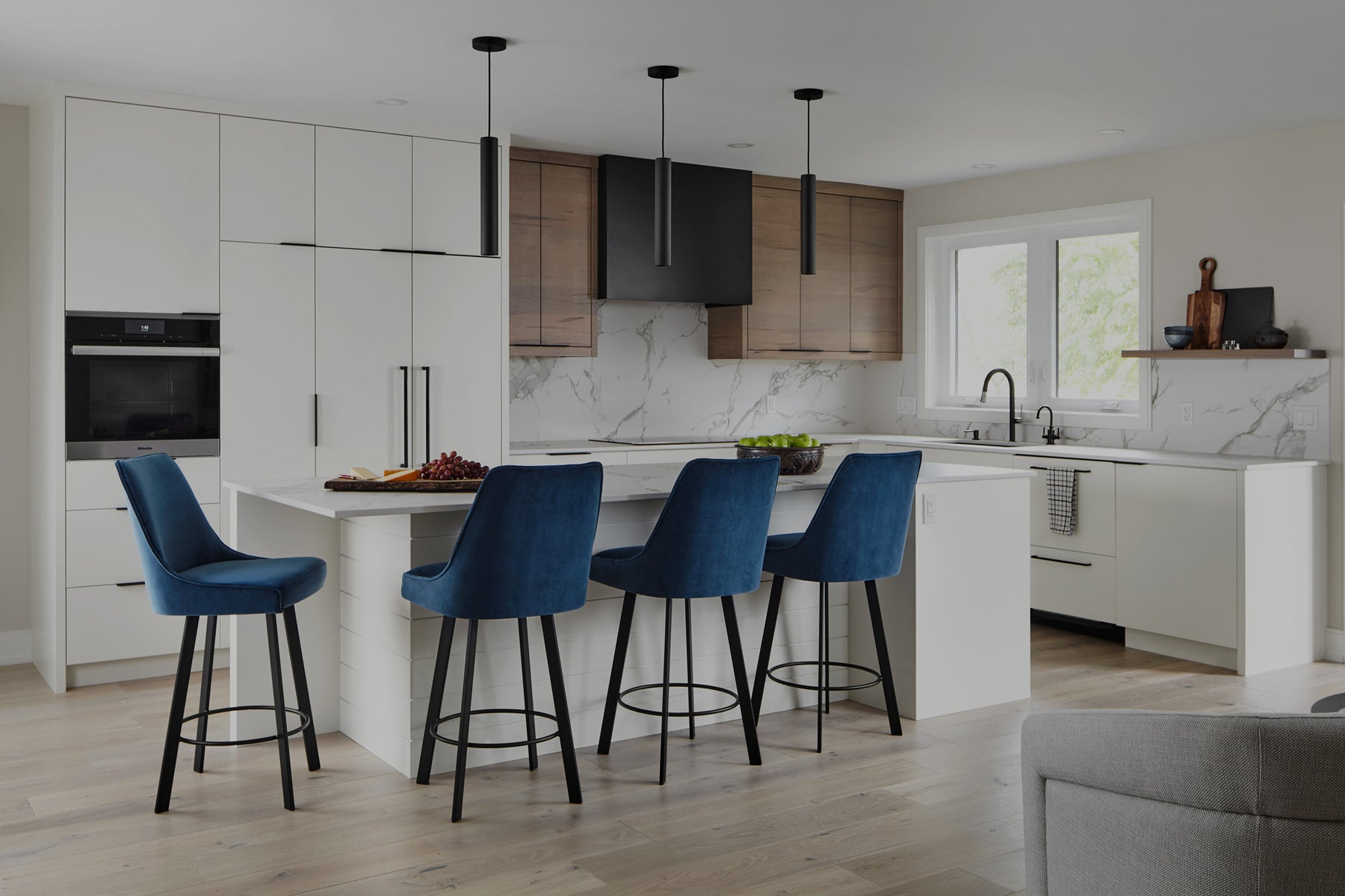 Modern kitchen renovation with white cabinets, contrasting blue fabric counter height stools, custom black hook and porcelain countertops white with grey veining.